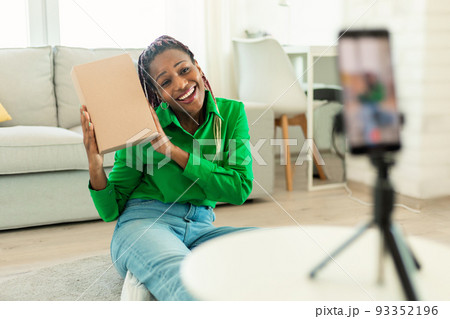 Happy black woman holding cardboard box and talking at smartphone camera on tripod Happy black woman holding cardboard box and talking at smartphone camera on tripod 93352196