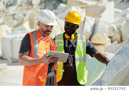 Two interracial male engineers in hardhats and uniform looking at tablet screen Two interracial male engineers in hardhats and uniform looking at tablet screen 93352742