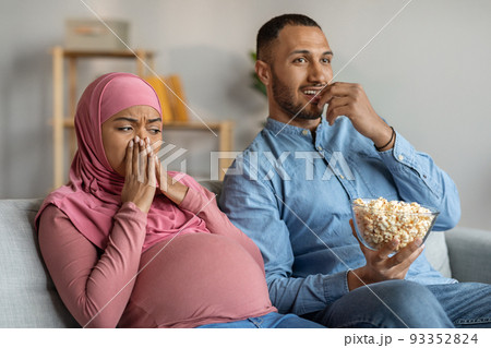 Black Pregnant Muslim Woman Feeling Disgusted While Husband Eating Popcorn At Home Black Pregnant Muslim Woman Feeling Disgusted While Husband Eating Popcorn At Home 93352824