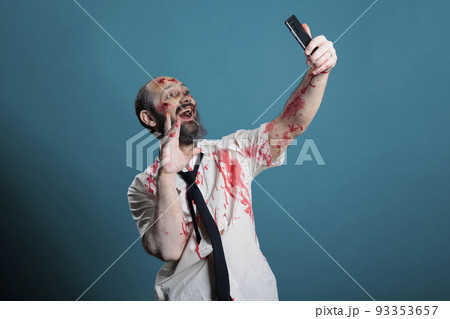 Apocalyptic corpse taking photo on smartphone, fooling around on camera and acting eerie. Spooky zombie with ugly wounds and bloody scars taking picture on mobile phone, brain eating. Apocalyptic corpse taking photo on smartphone, fooling around on camera and acting eerie. Spooky zombie with ugly wounds and bloody scars taking picture on mobile phone, brain eating. 93353657
