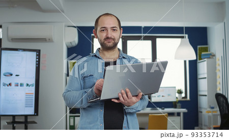 Portrait of casual business man holding laptop typing on keyboard and smiling at camera in busy modern office. Confident startup employee working relaxed in finance department of small company. 93353714