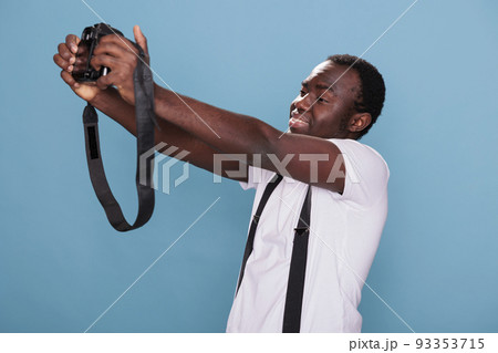 Smiling photography enthusiast taking picture of himself with modern DSLR camera while standing on blue background. Professional photographer having photo device while taking selfie photo. 93353715