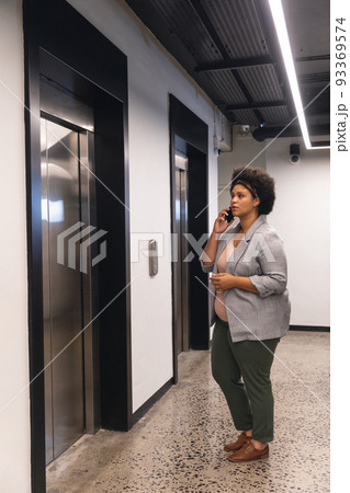 Mid adult biracial businesswoman talking on smartphone while waiting for elevator in office building 93369574