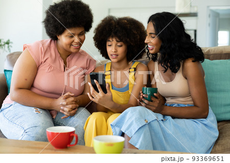 Biracial young woman using smartphone while sitting with female friends on sofa at home Biracial young woman using smartphone while sitting with female friends on sofa at home 93369651