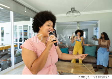 Happy biracial young woman with afro hair singing over microphone and friends dancing in background Happy biracial young woman with afro hair singing over microphone and friends dancing in background 93369657