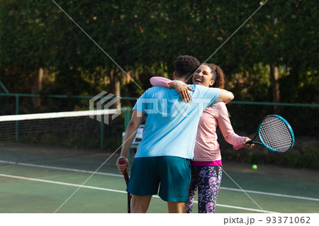 Smiling biracial couple playing tennis embracing on outdoor tennis court 93371062