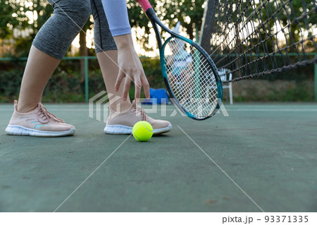 Low section of woman holding tennis racket picking up ball on outdoor tennis court, with copy space Low section of woman holding tennis racket picking up ball on outdoor tennis court, with copy space 93371335