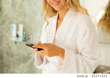 Happy caucasian woman wearing robe, using smartphone in bathroom 93371423