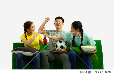 A group of young asian man and women holding football ball popcorn bowl sitting on green sofa cheering the soccer game at home on the white screen background. 93371626
