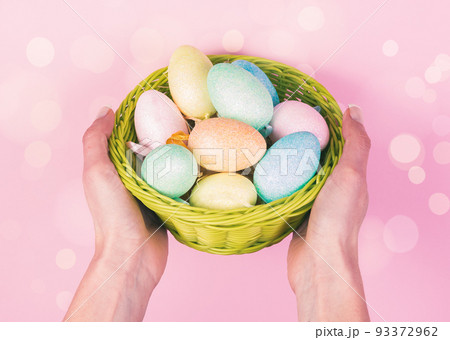 Woman hands holding wooden basket with colored ester eggs on pink background. Easter Concept 93372962