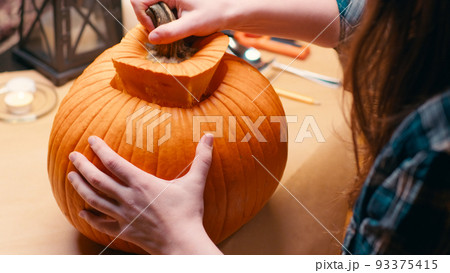 Preparing pumpkin for Halloween. Taking out lid and seeds. Woman sitting and carving halloween Jack O Lantern pumpkin at home for her family. 93375415