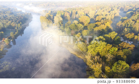 Aerial drone view flying over the river with calm reflective water surface and white fog mist and forest trees on the banks of the river on a sunny early autumn morning. Beautiful natural background. Aerial drone view flying over the river with calm reflective water surface and white fog mist and forest trees on the banks of the river on a sunny early autumn morning. Beautiful natural background. 93388690