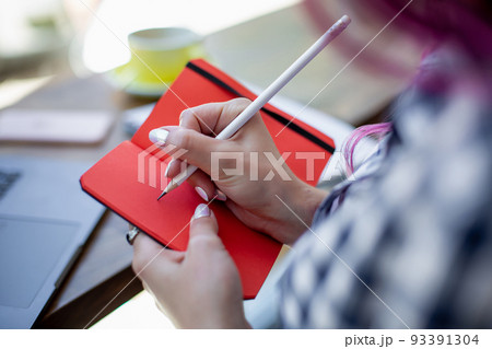 Positive female student with pink hair writing down funny story in notebook sitting in coffee shop with cup of cappuccino and laptop. 93391304