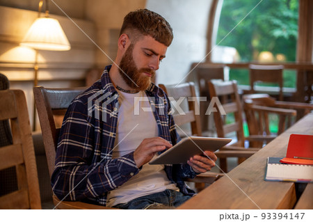 Freelancer dressed in casual outfit focus on reading news and looking on digital tablet while sitting in cozy urban cafe. 93394147