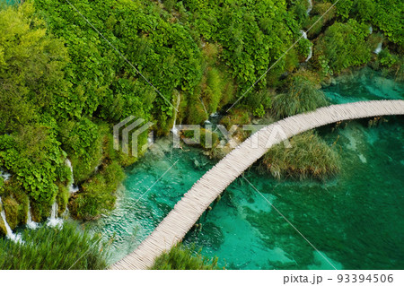 Hiking tourists walk on wooden footbridge surrounding lake with turquoise water in natural reserve. Majestic view of blue lake among picturesque landscape on Plitvice lakes upper view Hiking tourists walk on wooden footbridge surrounding lake with turquoise water in natural reserve. Majestic view of blue lake among picturesque landscape on Plitvice lakes upper view 93394506