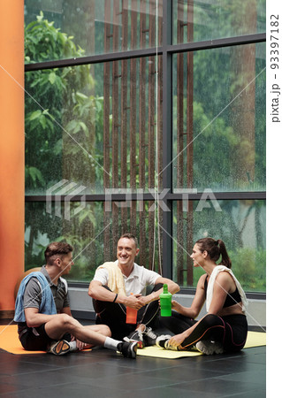 Positive young men and women drinking protein shakes when chatting after training in gym Positive young men and women drinking protein shakes when chatting after training in gym 93397182