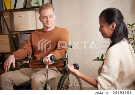 Young female caregiver showing man with disability how to exercise with dumbbells Young female caregiver showing man with disability how to exercise with dumbbells 93398103