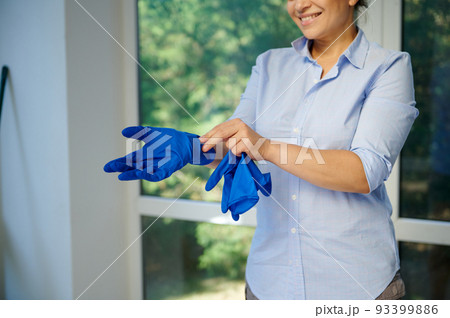 Close-up of charming young woman housewife, homemaker, maid putting on blue rubber work gloves to clean the house and wash windows during the spring general cleaning. Housekeeping. Household. Chores 93399886