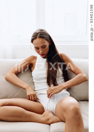 A beautiful woman poses on the couch at home in her home clothes in a white tank top and blue jeansHome lifestyle and body care about 93401596