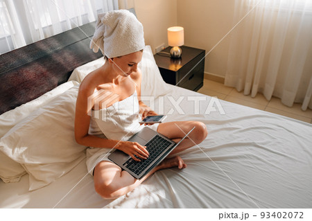 Happy caucasian woman using wireless laptop while resting on bed in bath towel. Female traveler enjoying time at comfy hotel room with modern device. Happy caucasian woman using wireless laptop while resting on bed in bath towel. Female traveler enjoying time at comfy hotel room with modern device. 93402072
