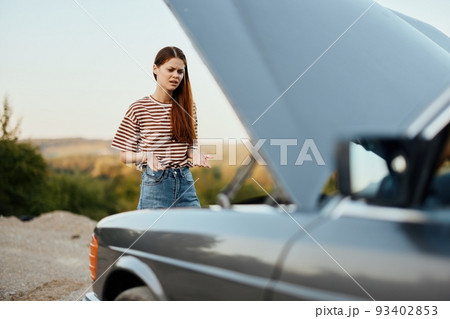 Woman traveler standing near the car with the hood open and looking for the cause of the car breakdown alone without men on the road in the countryside 93402853