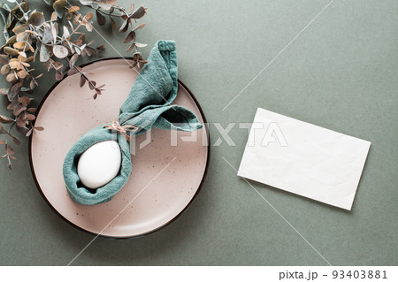 A white chicken egg is wrapped in a napkin in the form of a rabbit on a plate, a piece of paper for writing and a branch of eucalyptus on a green background. Eco friendly happy easter. Top view. 93403881