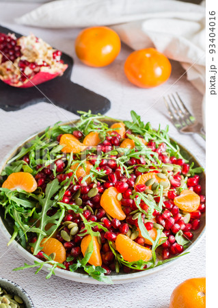 Fruit vitamin salad of pomegranate, tangerine, arugula and pumpkin seeds in a plate on the table. Organic vegetarian food. Vertical view. Close-up Fruit vitamin salad of pomegranate, tangerine, arugula and pumpkin seeds in a plate on the table. Organic vegetarian food. Vertical view. Close-up 93404016