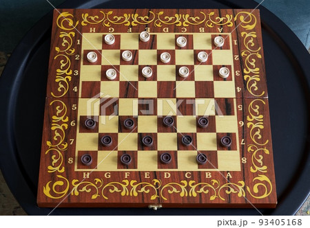 Board game of checkers - a wooden board with placed checkers stands on a round black metal table in the living room. Board game of checkers - a wooden board with placed checkers stands on a round black metal table in the living room. 93405168