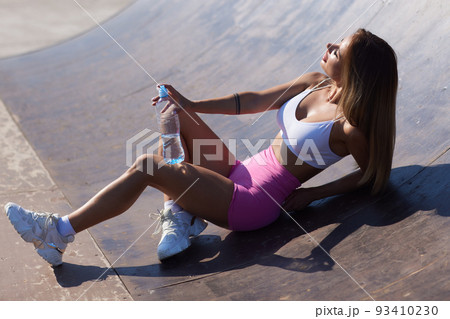 Young tired fit woman in sports clothes is resting leaning against the wall and drinking water in the city. 93410230