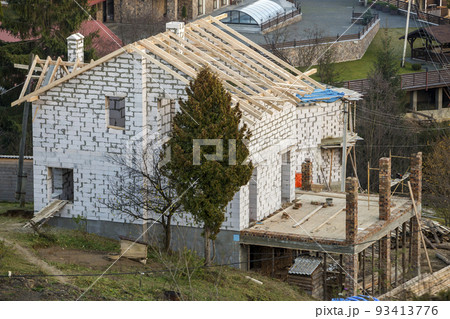 Two-storied cottage under construction. Walls of hollow foam insulation blocksand with window openings,roof beams frame and high chimneys, roofing underlayment. 93413776