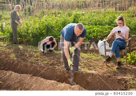 Father digs trench while the rest of the family is engaged in garden Father digs trench while the rest of the family is engaged in garden 93415230