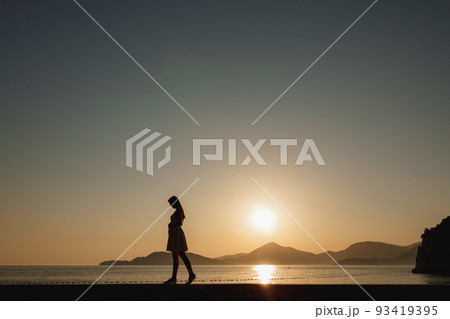 A pregnant woman walks on the seashore at sunset and gently strokes her belly, behind her are mountains and the setting sun  93419395