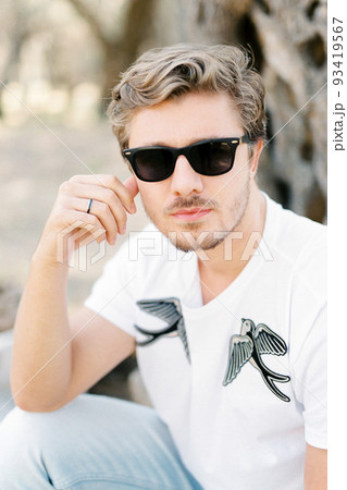 Young man in sunglasses sits touching his cheek with fingers Young man in sunglasses sits touching his cheek with fingers 93419567
