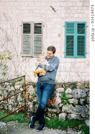 Dad with a baby in his arms stands at the stone wall of the house leaning on the foundation 93419974
