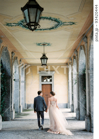Bride and groom walk holding hands along the terrace of an old villa. Back view 93420444