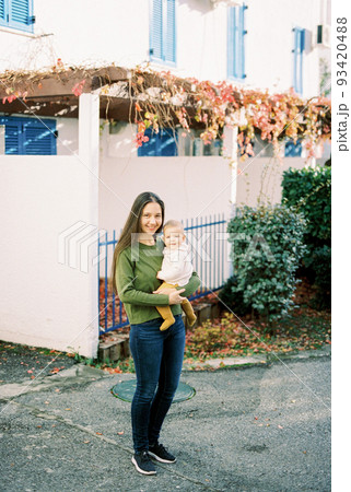 Mom with a baby in her arms stands in the yard near the house 93420488