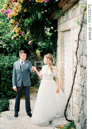 Bride and groom stand near a stone wall under a flowering tree Bride and groom stand near a stone wall under a flowering tree 93420579