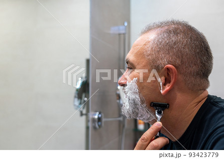 Middle aged handsom man trimming his beard and mustache standing in bathroom and looking in a mirror Middle aged handsom man trimming his beard and mustache standing in bathroom and looking in a mirror 93423779
