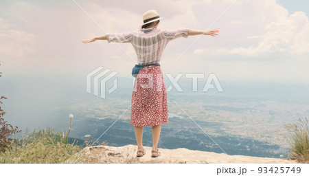 Woman traveler flaps arms like wings on mountain top. Young woman in hat opens arms wide waving like bird's wings over Tira city, Albania. Red dress happy girl blowing wind. Dream, travel concept 93425749