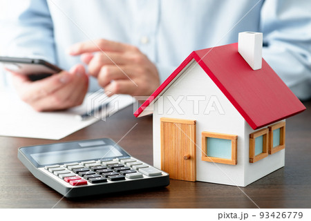 テーブルの上のに置かれた電卓と住宅模型。スマートフォンを使って調べる男性。 93426779