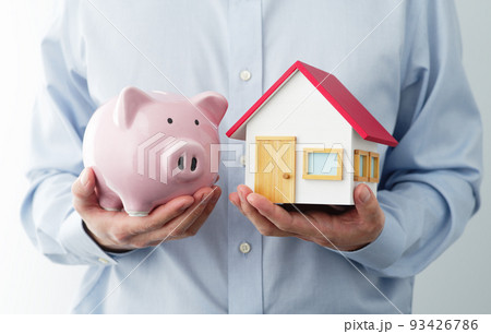 ブタの貯金箱と家の模型を持つ男性。家計の節約のイメージ。 93426786