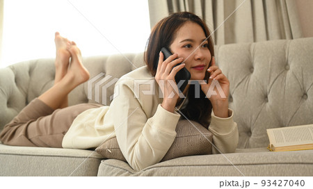 Beautiful young woman lying on couch talking on mobile phone, spending leisure time in cozy winter or autumn weekend at home 93427040