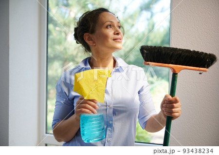 Close-up portrait of a Latin American pretty woman, housewife in blue casual shirt, looking aside, holding a broom and rag with detergent in her hands. Housekeeping and spring cleaning service concept Close-up portrait of a Latin American pretty woman, housewife in blue casual shirt, looking aside, holding a broom and rag with detergent in her hands. Housekeeping and spring cleaning service concept 93438234