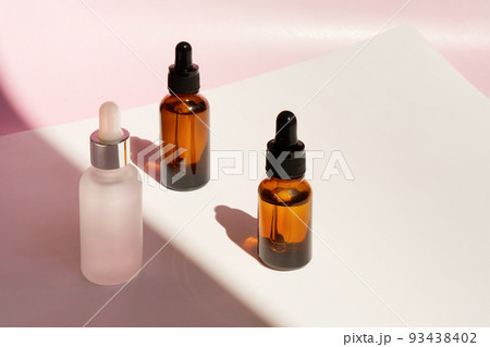 Three transparent amber and frozen glass dropper bottles with liquid inside on pink background in the sunlight. Pipette with fluid hyaluronic acid, serum, retinol. Cosmetics, healthcare, beauty  93438402