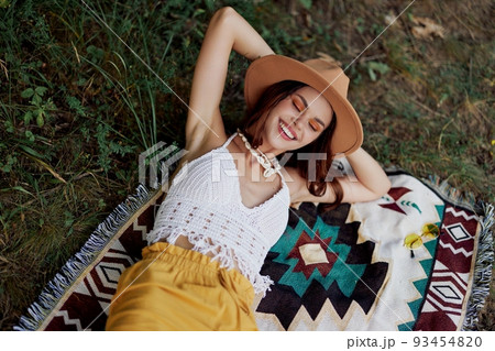 A woman in eco-friendly clothing in a hippie look is lying on a colored plaid smiling and looking at an autumn sunset in nature in the park. Lifestyle on a journey of harmony with the world 93454820