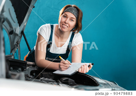 Portrait of young smiling mechanic woman in coveralls signs documents leaning at the car. The concept of woman's equality and works at auto repair shop Portrait of young smiling mechanic woman in coveralls signs documents leaning at the car. The concept of woman's equality and works at auto repair shop 93479883