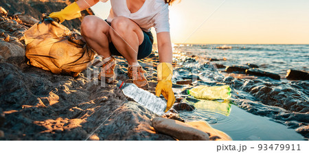 Global environmental pollution banner. A female volunteer sitting collects plastic bottles on the ocean shore. Cleaning of the coastal zone. Copy space 93479911