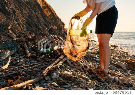 Woman collects rubbish in a plastic bag. In the background the sea and wild beach. Close up of woman's legs. Low angle view. The concept of coastal zone cleaning 93479912