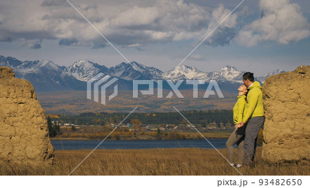 Man and woman in yellow green sportswear. Lovely couple of travelers hug and kiss near old stone enjoying highland landscape. Man and woman in yellow green sportswear. Lovely couple of travelers hug and kiss near old stone enjoying highland landscape. 93482650