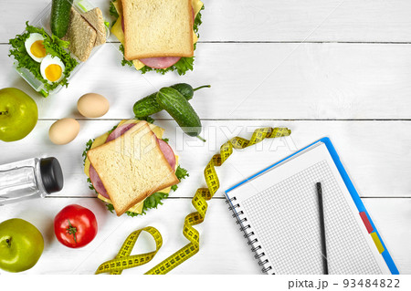 Top view planning notebook with copy space and healthy breakfast. Sandwich with ham and cheese, eggs, cucumber, tomato and bottle of water on white wooden table. 93484822